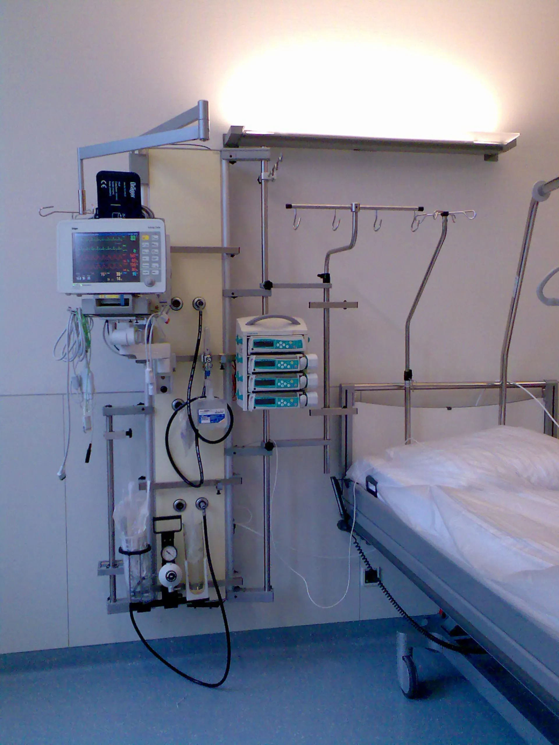 Krankenhauszimmer mit medizinischer Wandkonsole, Monitor, Infusionspumpen, Sauerstoffanschlüssen und einem Bett mit weißer Bettdecke